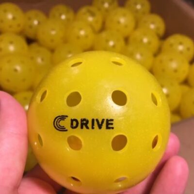 The USAPA Approved C DRIVE Pickleball up Close with a box of yellow C DRIVE Pickleballs in the background