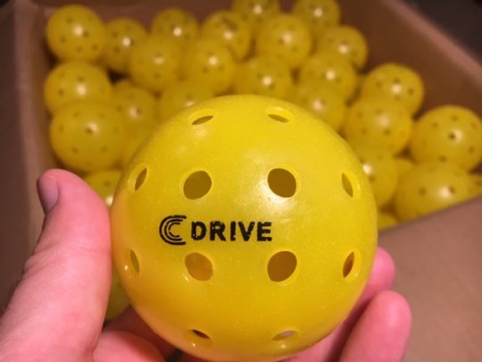 The USAPA Approved C DRIVE Pickleball up Close with a box of yellow C DRIVE Pickleballs in the background