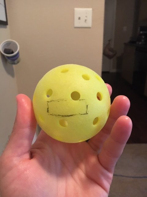 Calvin Keeney's hand holding a Yellow pickleball with a 1 inch by 3 inch blank box on it where the C DRIVE pickleball logo will eventually go