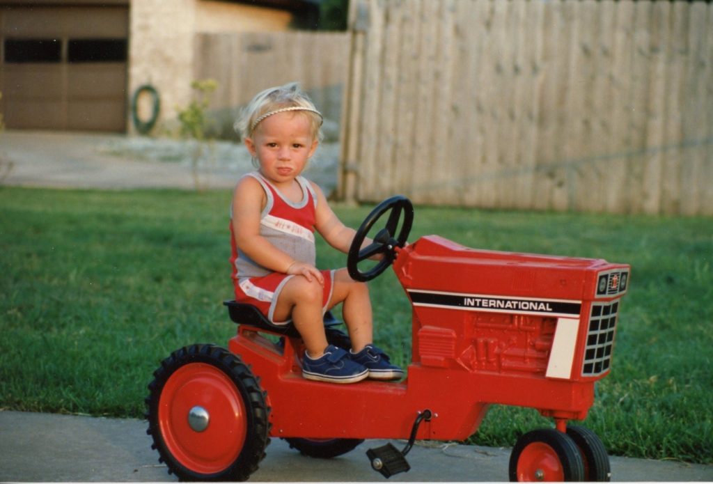 Calvin Keeney sitting on a small red tractor while driving it and looking at the camera. He was about 4 years old