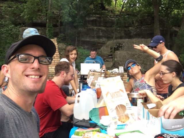 A group of rock climbers outdoors at a picnic table eating food together. Calvin Keeney sitting next to Brian Adrian with Alexa waving at the Camera while Jeremy cooks. Along with 3 other people